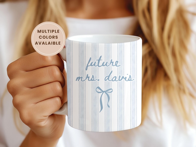 Future Mrs. personalized mug with blue coquette bow design and soft vertical stripes. Bride-to-be engagement gift featuring custom last name text on a ceramic coffee mug. Aesthetic bridal shower gift idea shown being held in hand, perfect for fiancées, wedding planning, and proposal celebrations. Customizable colors available for a unique, thoughtful gift.