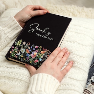Puede incluir: Un diario negro con el texto "Sarah's New Chapter" en escritura blanca. La parte inferior del diario presenta un dise&ntilde;o floral colorido. El diario est&aacute; abierto y sostenido por una persona.