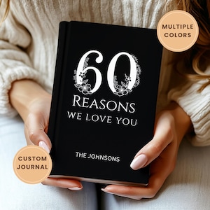May include: A black journal with the text "60 REASONS WE LOVE YOU" in white. The number 60 is decorated with floral accents. The journal also includes the name "THE JOHNSONS" at the bottom. The journal is held in someone's hands.