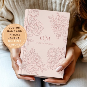 May include: A pale pink journal with floral line art and the initials "OM" and the name "Olivia Mason". The journal cover features a peony design. Text on the journal reads "CUSTOM NAME AND INITIALS JOURNAL MULTIPLE COLORS".