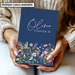 Puede incluir: Un diario azul marino con el nombre "Olivia" y "Capítulo 30" en escritura blanca. El borde inferior presenta un diseño floral colorido. El texto "Multiple Colors Available" está en la esquina superior izquierda.