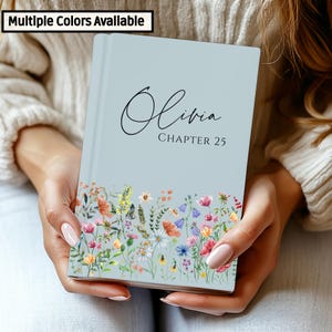 May include: A light blue journal with the name "Olivia" and "Chapter 25" printed on the cover. The bottom of the journal features a colorful floral design. The text "Multiple Colors Available" is in the top left corner.