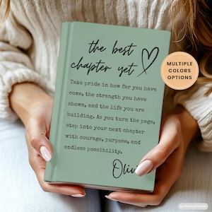 May include: A sage green hardcover journal featuring the black printed text "the best chapter yet" with a heart icon, followed by an inspirational paragraph and the name "Olivia" at the bottom. A circular badge indicates multiple color options.