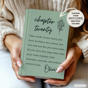 May include: A sage green hardcover book with black text that reads chapter twenty, followed by an inspirational quote about personal growth and the name Olivia. A small black line drawing of a flower is featured, with a circular label above.
