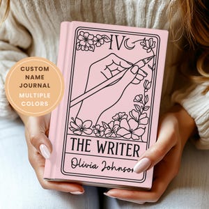 Puede incluir: Un diario rosa personalizado con una ilustración negra de una mano sosteniendo un bolígrafo, rodeada de diseños florales. El texto "THE WRITER" y un nombre están debajo. El diario se sostiene con ambas manos.
