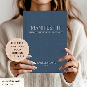 Puede incluir: Un diario azul con texto plateado que dice "MANIFEST IT". El texto "THINK IT • BELIEVE IT • BECOME IT" está debajo. El nombre "ISABELLA DAVIS" y el año "2026" están impresos en la parte inferior. El diario es sostenido por una persona.