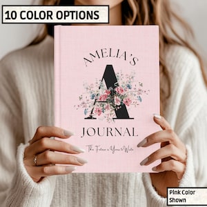 May include: A pink journal with the name "Amelia's" and the word "Journal" printed on the cover. A large black "A" is decorated with a floral arrangement. The text "The Future is Yours to Write" is below the word "Journal". The text "10 COLOR OPTIONS" is in a black box.