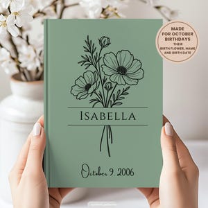 May include: A sage green hardback book featuring a black line art illustration of a flower bouquet. The name ISABELLA is printed in black capital letters across the centre, with the date October 9, 2006, below. A circular badge reads MADE FOR OCTOBER BIRTHDAYS.