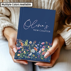 Könnte beinhalten: Ein marineblaues Notizbuch mit dem Namen "Olivia's" und den Worten "New Chapter" in weißer Schrift. Der untere Teil des Notizbuchs zeigt ein farbenfrohes Blumendesign mit verschiedenen Blumen. "Multiple Colors Available" befindet sich oben links.