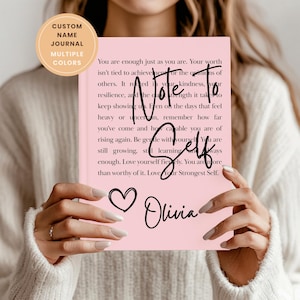 May include: A pink journal with the text "Note to Self" and the name "Olivia" in cursive. The journal has a heart graphic and a circular sticker that says "Custom Name Journal Multiple Colors". The journal is held by a person.