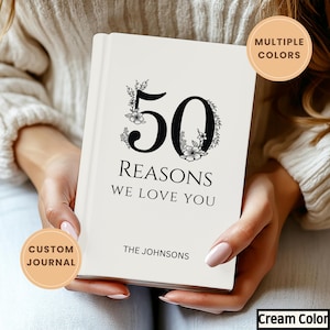 Puede incluir: Diario de color crema con el texto "50 REASONS WE LOVE YOU" en negro. El número 50 está decorado con detalles florales. El diario está personalizado con el nombre "THE JOHNSONS" en la parte inferior. El diario es un diario personalizado.