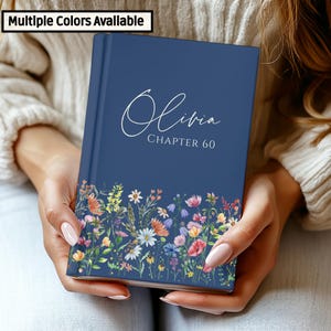 May include: A navy blue book with the name "Olivia" and "Chapter 60" in white script. The bottom of the book features a floral design with colorful flowers. The top left corner of the book has the text "Multiple Colors Available".