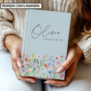May include: A pale blue book with the name "Olivia" and "CHAPTER 21" printed on the cover. The bottom of the book features a floral design with colourful flowers. The text "Multiple Colors Available" is in the top left corner.