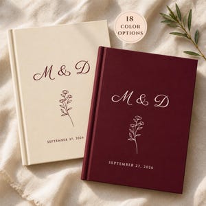 Op de afbeelding: Twee hardcover journals, één crème en één bordeaux, met de initialen "M & D" en een bloemenillustratie. De datum "27 september 2026" staat onderaan gedrukt. Een cirkelvormige afbeelding geeft "18 COLOR OPTIONS" aan.