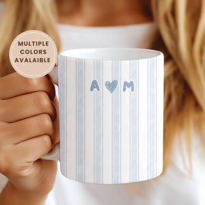 Pode incluir: Caneca de cerâmica branca com um padrão de riscas azul claro. A caneca apresenta as letras "A" e "M" com um símbolo de coração no meio. A caneca é segurada por uma pessoa. O texto "MULTIPLE COLORS AVALAIBLE" está num autocolante redondo.