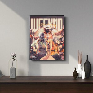 May include: A black framed poster with a colorful image of The Weeknd. The poster features the text "WEEKND" in large, stylized letters. The image shows The Weeknd wearing a mask and a white shirt, with other figures in the background.