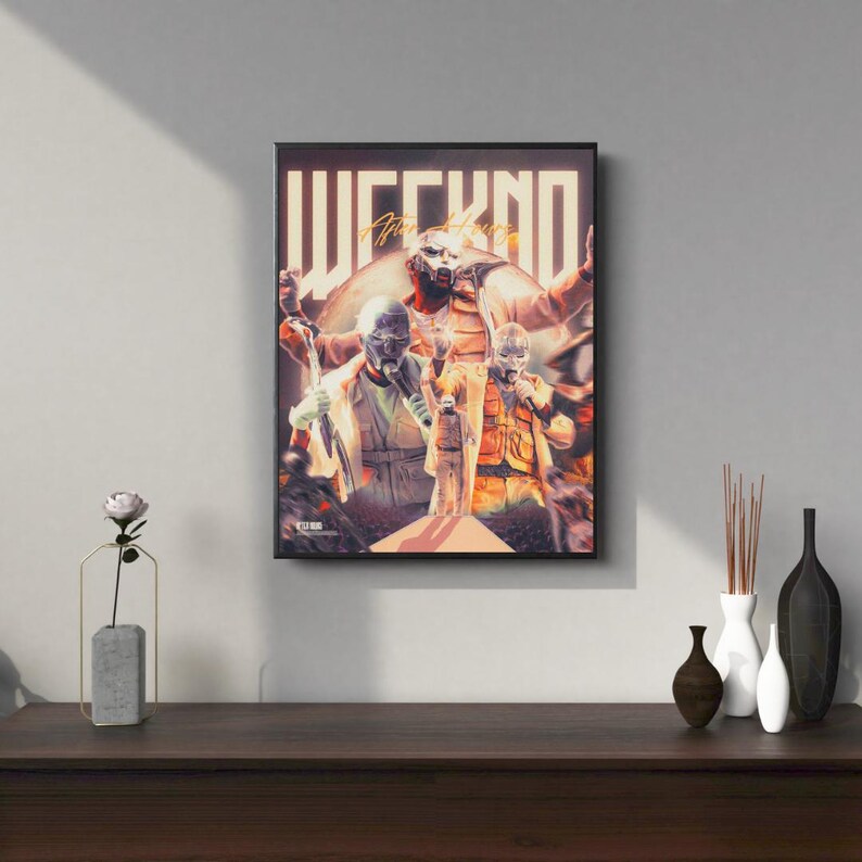 May include: A black framed poster with a colorful image of The Weeknd. The poster features the text "WEEKND" in large, stylized letters. The image shows The Weeknd wearing a mask and a white shirt, with other figures in the background.