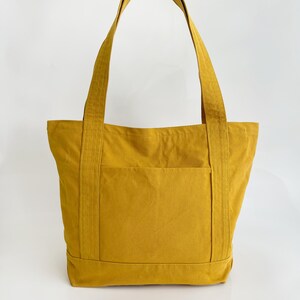 May include: Mustard yellow tote bag with a front pocket and strong handles. The bag is made of a durable fabric, designed for carrying items. The tote bag is a solid colour and has a simple, classic design.