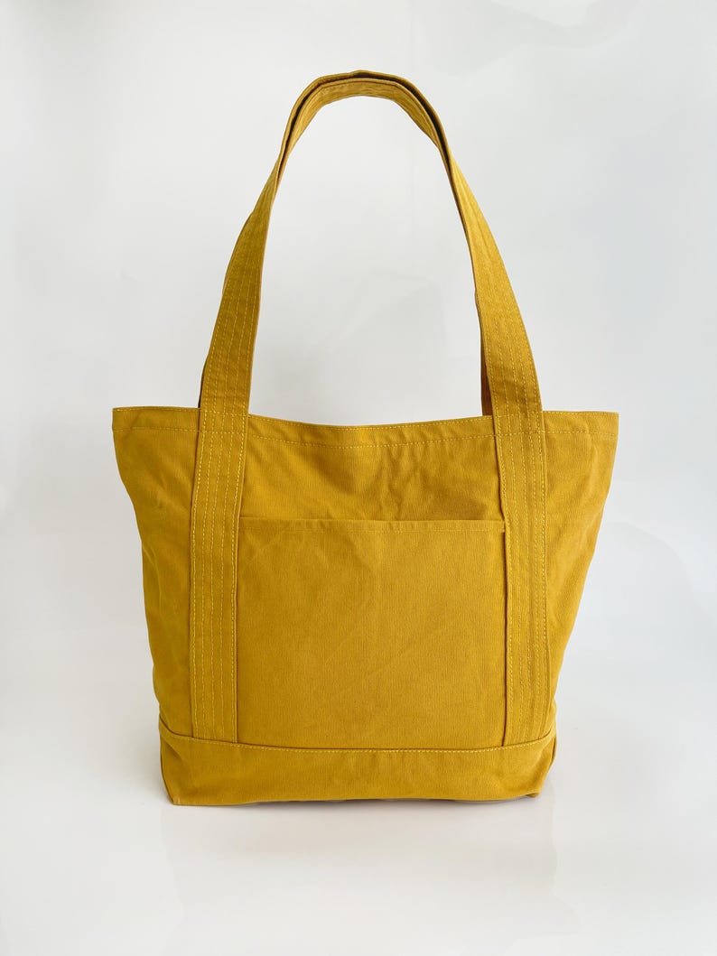 May include: Mustard yellow tote bag with a front pocket and strong handles. The bag is made of a durable fabric, designed for carrying items. The tote bag is a solid colour and has a simple, classic design.