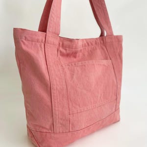 May include: A dusty rose tote bag made of sturdy fabric. The bag features a large front pocket and two long straps for carrying over the shoulder. The bag is designed with a rectangular shape and is suitable for everyday use.