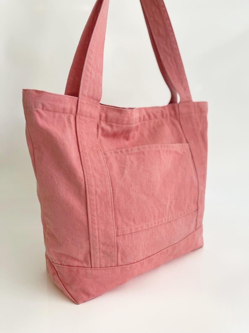 May include: A dusty rose tote bag made of sturdy fabric. The bag features a large front pocket and two long straps for carrying over the shoulder. The bag is designed with a rectangular shape and is suitable for everyday use.