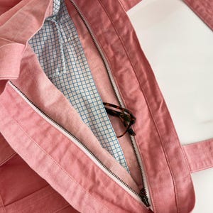 May include: A pink denim tote bag with a zippered main compartment. The bag has sturdy straps and a blue and white grid pattern lining. Tortoiseshell eyeglasses are visible inside the bag.