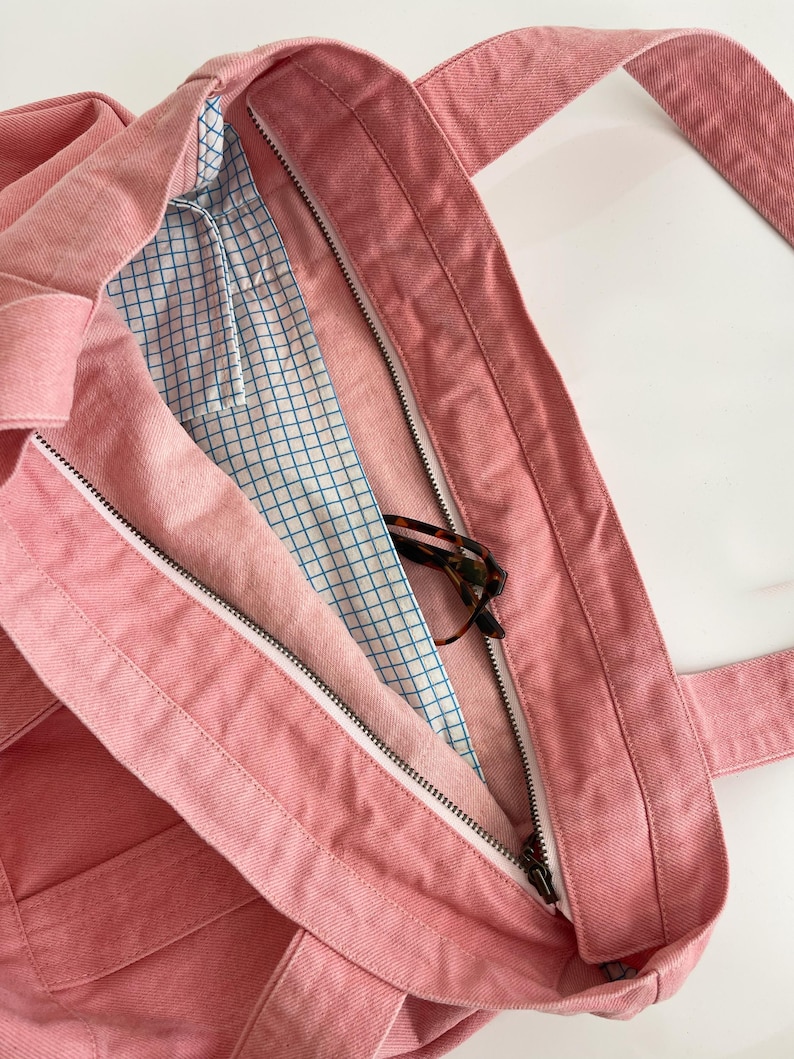 May include: A pink denim tote bag with a zippered main compartment. The bag has sturdy straps and a blue and white grid pattern lining. Tortoiseshell eyeglasses are visible inside the bag.