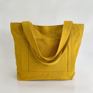 May include: Mustard yellow tote bag with two long straps and a front pocket. The bag is made of a sturdy fabric and has a rectangular shape. The straps are sewn to the bag's body. The bag is designed for carrying items.