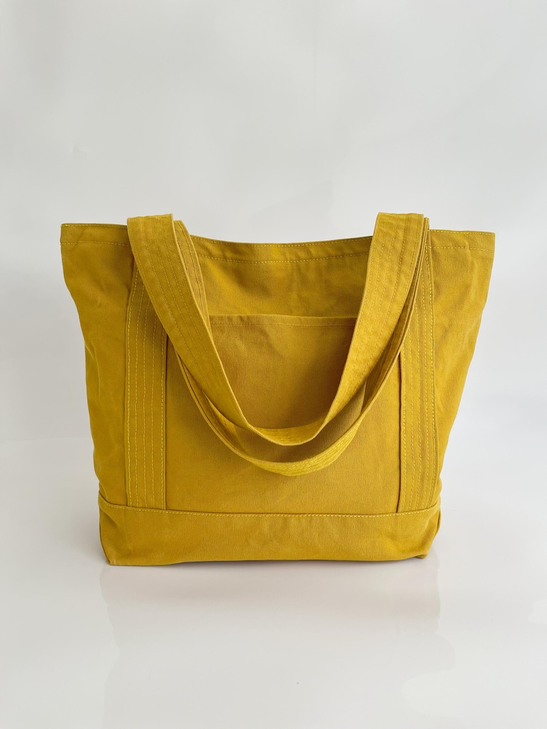 May include: Mustard yellow tote bag with two long straps and a front pocket. The bag is made of a sturdy fabric and has a rectangular shape. The straps are sewn to the bag's body. The bag is designed for carrying items.