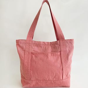 May include: A dusty rose tote bag with a rectangular shape and a front pocket. The bag has two long handles and is made of a sturdy fabric. The background is plain white.