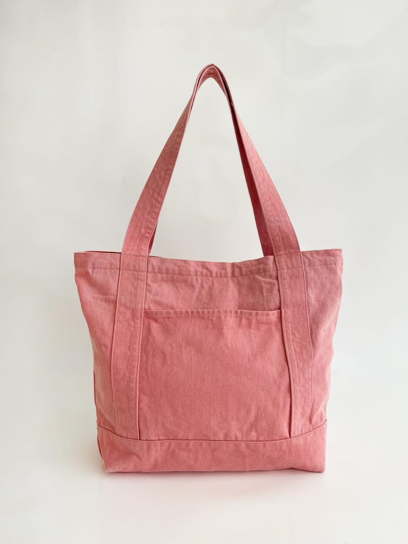 May include: A dusty rose tote bag with a rectangular shape and a front pocket. The bag has two long handles and is made of a sturdy fabric. The background is plain white.