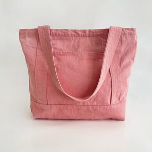 May include: A dusty rose tote bag made of sturdy fabric. The bag has two long straps and a rectangular shape with a pocket on the front. The bag is designed for carrying items and has a simple, minimalist style.