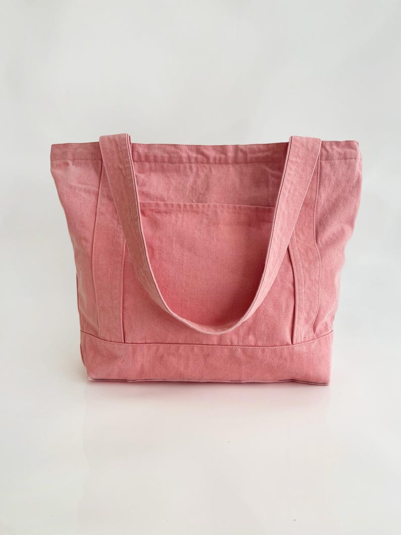 May include: A dusty rose tote bag made of sturdy fabric. The bag has two long straps and a rectangular shape with a pocket on the front. The bag is designed for carrying items and has a simple, minimalist style.