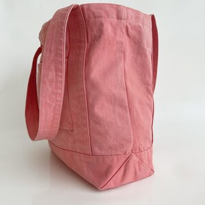 May include: A pink tote bag made of sturdy fabric. The bag has long straps and a rectangular shape with a flat bottom. The bag is a solid colour and appears to be empty.