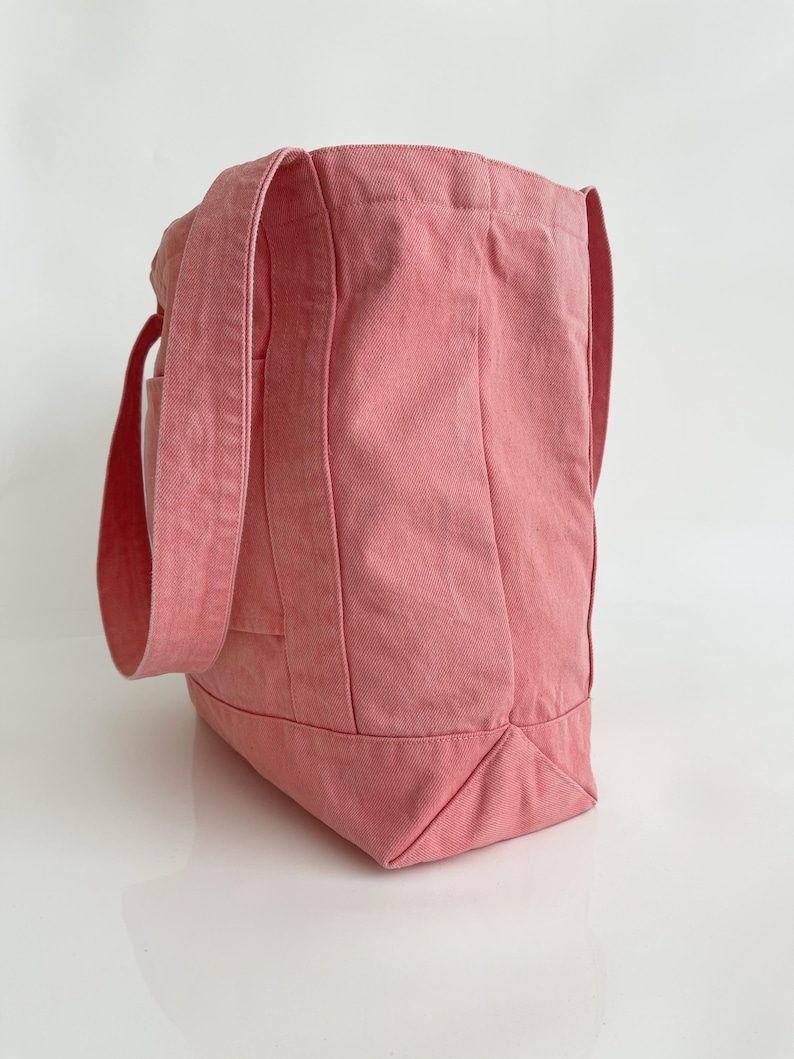 May include: A pink tote bag made of sturdy fabric. The bag has long straps and a rectangular shape with a flat bottom. The bag is a solid colour and appears to be empty.