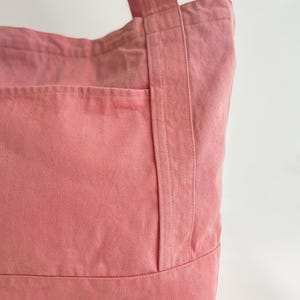 May include: A close-up of a pink tote bag made of sturdy fabric. The bag has a large front pocket and two handles. The stitching is visible, and the bag appears to be well-constructed. The colour is a soft, muted pink.