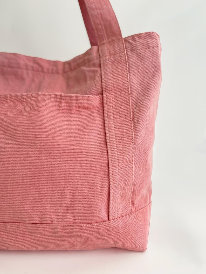 May include: A close-up of a pink tote bag made of sturdy fabric. The bag has a large front pocket and two handles. The stitching is visible, and the bag appears to be well-constructed. The colour is a soft, muted pink.