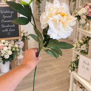 May include: A single artificial flower stem featuring one large, fully bloomed cream-coloured peony with a yellow centre and one smaller, closed white peony bud. The stem includes several dark green leaves attached to a thin, straight green stalk.