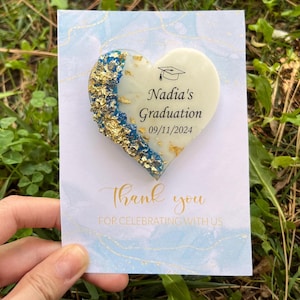May include: A heart-shaped graduation keepsake with the text "Nadia's Graduation 09/11/2024" and a graduation cap icon. The heart is decorated with gold and blue accents, and is on a card that says "Thank you".