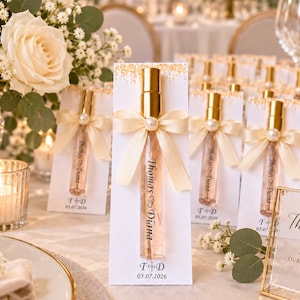 Puede incluir: Un frasco de perfume de vidrio transparente con tapa dorada, sujeto a una tarjeta rectangular blanca con una cinta color crema y una perla artificial. La tarjeta presenta detalles dorados, el texto Thomas & Diana y la fecha 05.07.2026.