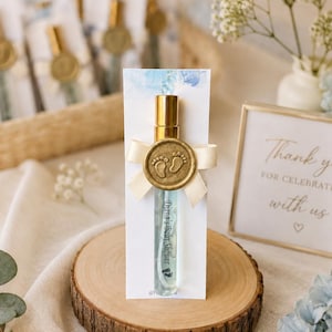 Op de afbeelding: Een kleine glazen sprayfles met een gouden dop, bevestigd aan een witte kaart met blauwe aquarelaccenten. De kaart bevat de tekst Diana's Baby Shower en een gouden waszegel met babyvoetafdrukken, vastgemaakt met een crèmekleurig lint.