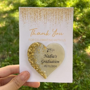 May include: A heart-shaped graduation favor with gold leaf accents, personalized with "Nadia's Graduation" and the date 09/11/2024. The card reads "Thank You For Celebrating With Us."