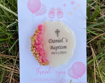 May include: A circular white resin ornament with a textured pink and gold leaf edge, featuring a black cross icon and the text Daniel's Baptism 09/11/2024. It is mounted on a white card with pink balloons, baby shoes, and the text Thank you for celebrating with us.
