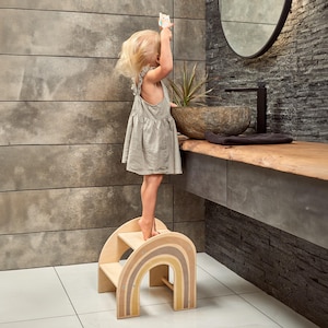 Può includere: Uno sgabello a forma di arcobaleno in legno con due gradini. Lo sgabello è dipinto in tonalità di marrone e giallo. Un bambino piccolo è in piedi sul gradino superiore.