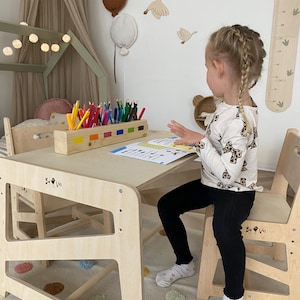 Könnte beinhalten: Ein Kindertisch- und Stuhlset aus hellem Holz. Der Tisch hat eine rechteckige Oberseite und ein unteres Regal. Auf dem Tisch befindet sich ein Stifthalter aus Holz mit Buntstiften. Der Stuhl ist ebenfalls aus hellem Holz. Der Raum hat Wanddekorationen.