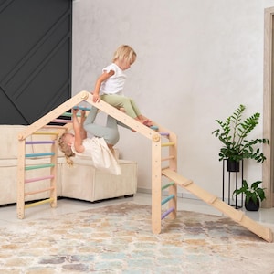 Może przedstawiać: Drewniana konstrukcja do zabawy w pomieszczeniu z ścianą wspinaczkową, zjeżdżalnią i drążkiem do ćwiczeń. Dwoje dzieci bawi się na konstrukcji. Ściana wspinaczkowa ma kolorowe szczeble, a zjeżdżalnia jest drewniana.