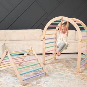 Può includere: Un telaio per arrampicata in legno con pioli color arcobaleno. Un bambino è appeso al piolo superiore del telaio. C'è un telaio per arrampicata in legno più piccolo in primo piano.