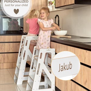 Può includere: Due sgabelli bianchi in legno per bambini, con una targhetta con il nome "Jakub". Gli sgabelli sono in una cucina. È visibile anche il testo "PERSONALIZED Just for your child".