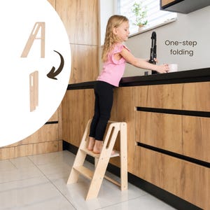 Può includere: Uno sgabello a gradini in legno chiaro con due gradini e maniglie laterali. Un grafico inserito mostra lo sgabello in posizione aperta e piegata con il testo One-step folding. Lo sgabello è realizzato in legno chiaro con una finitura liscia.