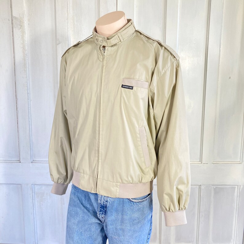 May include: A beige bomber jacket with a zipper closure and a brown logo patch that reads "Members Only" on the left chest.